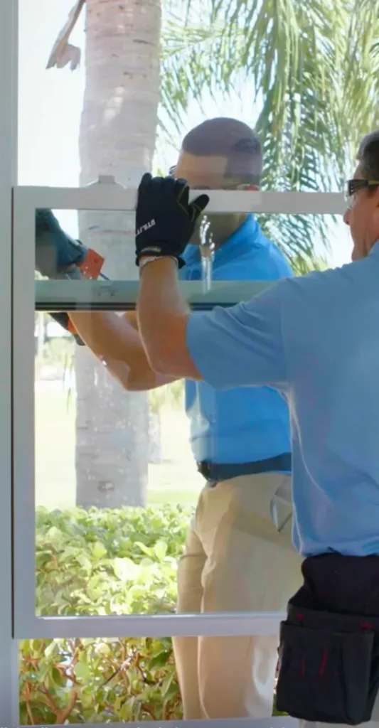 Workers installing a window pane in a bright outdoor setting with palm trees in the background.