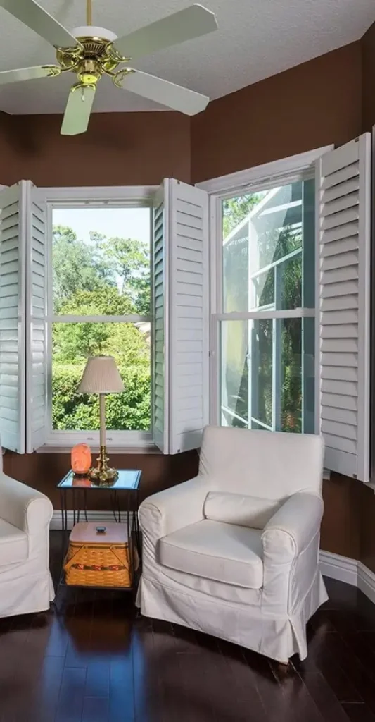 Cozy sitting area with white armchairs beside a window with open shutters overlooking greenery.